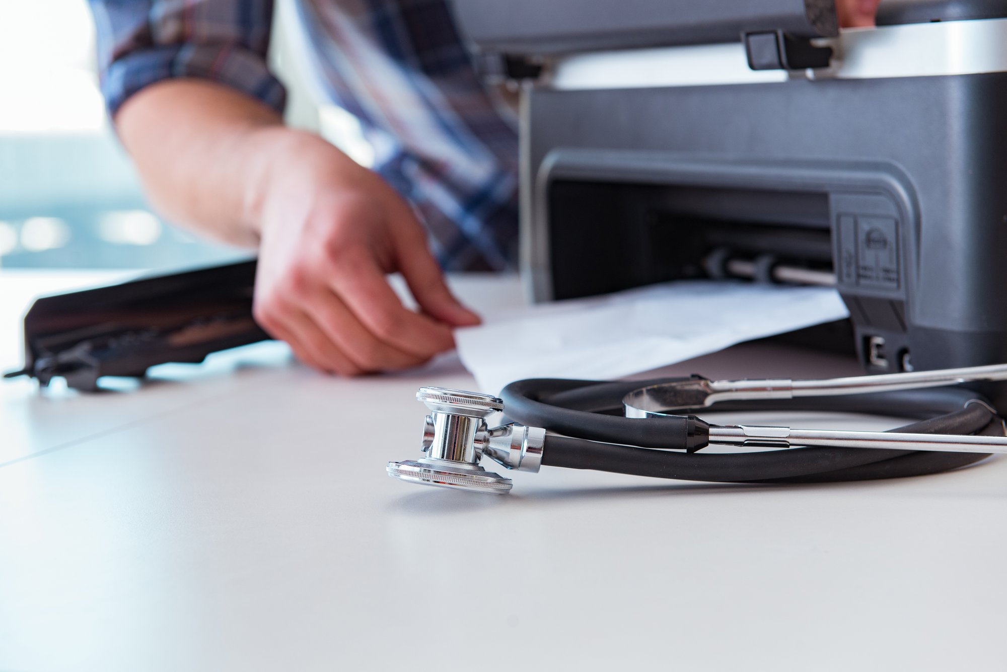 a desk with a stethoscope and a printer. a hand is retrieving a paper in the printer.